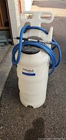 Full image of white BioSafe portable tank with blue hose and attached white spray nozzle, showing tank body, handle, hose coiled and nozzle held in hose loops.