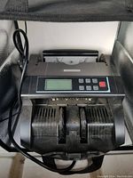 Front view of the black electronic bill counter showing LCD screen, control buttons, bill feeding slots, and attached power cord.