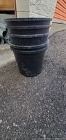 Stack of five black plastic plant pots against a wall and pavement background.