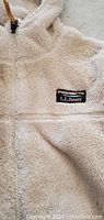 Close-up photo of the front of the hoodie showing the LL Bean logo patch on the chest and the plush cream-colored polyester fabric texture.