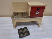 Vintage metal toy stove with cream-colored body and red oven door featuring a black knob and temperature gauge. Stove top has circular ventilation holes. Muffin pan with four slots positioned in front.