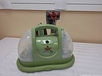 Full front view of Bissell Little Green Carpet Cleaner showing green body, transparent water tanks, coiled green hose, and brush attachment behind unit.