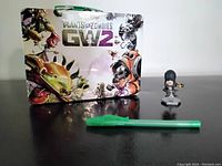 Plants vs Zombies Garden Warfare 2 tin lunchbox with colorful character artwork and green handle, alongside a small Assassins Creed mini figurine on a hexagonal base.