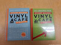 Two CD cases from the Vinyl Cafe series by Stuart McLean displayed side by side on a wooden surface. The left case has a blue background with orange and black text and the right case has a green background with red and black text.