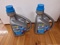 Two gray plastic containers of Alaskan Ice Preventer with blue caps and built-in handles on a wooden floor.