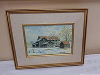 Framed oil painting showing rustic barns in snow with visible artist signature.