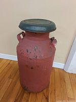 Front and side view of a 24-inch tall red painted metal milk can with a green metal lid on a hardwood floor against a beige wall.