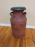 Front view of the painted milk can showing red painted body with rust and blue lid on hardwood floor.