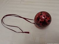 Photo showing red and black glass friendship ball ornament with long red ribbon on white surface, highlighting size and condition.