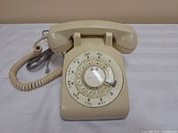 Front view showing rotary dial, numbers, letters, and handset on top of beige vintage telephone base.