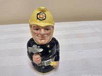 Front view of the fireman figurine showing facial features, yellow helmet with crest, black uniform, and items in hands.