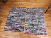 Full view of two rectangular handwoven rag rugs laid flat on wood floor showing multicolored stripes and fringed edges.