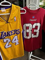 Photo showing front of yellow Los Angeles Lakers basketball jersey with number 24 and front of red Buccaneers T-shirt with number 83 and logo
