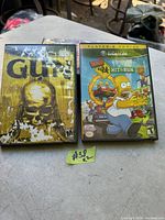 Photo showing two Nintendo GameCube game cases: Gun and The Simpsons Hit & Run, placed on a gray surface outdoors.
