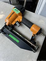 Close-up photo of an orange Bostitch pneumatic nail gun, showing the label, grip, and nail magazine.