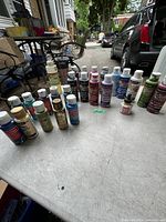 Photo showing a variety of acrylic paint bottles of different colors and brands arranged on a table outdoors.