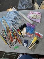 Top down view of the large collection of varied artist brushes arranged on a grey surface, including metal and wooden handled brushes of various sizes, also showing some packaged brushes and foam stencil brushes.
