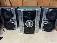 Photo shows Sony stereo system placed on a surface, featuring the central unit with dual cassette players, CD player, and AM/FM radio controls along with two matching Sony speakers on either side.