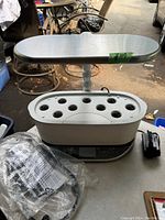 Full view of AeroGarden Bounty Elite Artisan indoor garden system with adjustable light panel and seed pod openings, placed on a table with power adapter and plastic bag beside.