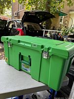 Green Sterilite heavy duty storage tote placed on a white table outdoors, front and side view showing metal latch closures and handles.