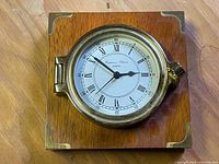 Front view of the Captain's Choice quartz clock on a polished wooden square base with brass corner accents, showing the clock face with Roman numerals.