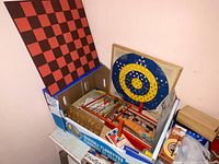 Photo showing large wooden checkerboard, dartboard, and several boxed games in a cardboard box.