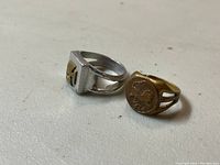 Two rings side by side on a light surface, showing the silver tone ring with square top and iron cross design, and the gold tone ring with a round top and lion emblem.