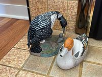 Overall view of duck and loon figurines together on tiled floor showing color and detail.