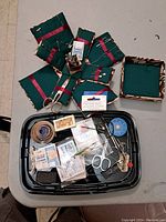 Full view of fabric needle holders, scissors, measuring tapes, needle packets, and sewing accessories on a black tray and one fabric-covered box for storage.