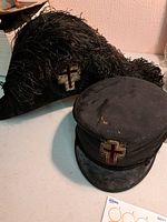 Two vintage Masonic hats displayed on a flat surface: a black feathered tricorne and a black Masonic cap with cross emblems.