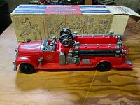 Front side view of the red vintage firetruck toy with attached hoses, ladder, and firemen figures, displayed on wooden surface with original box behind it.