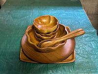 Top view of stacked wooden salad bowl set showing three small flower-shaped bowls inside a larger flower-shaped bowl, two wooden serving spoons on the side, and underneath a round tray and a square tray visible.