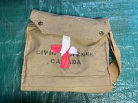 Front view of the olive drab canvas bag with flap closure showing 'CIVIL DEFENSE CANADA' stencil and painted red cross on front.