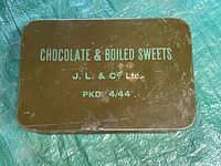 Top view of rectangular brown metal tin with blue text on lid 'CHOCOLATE & BOILED SWEETS, J.L. & Co Ltd, PKD 4/44'. Visible scratches and wear marks.
