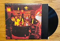 Photo of the front album cover showing band members around a table in a wood-paneled room with warm lighting.