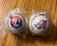 Two baseballs side by side on wood floor, one with Montreal Expos logo and one with Anaheim Angels logo, each wrapped in clear plastic.