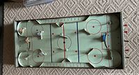 Full view of the rectangular metal table hockey game board showing painted rink with two goals and six player figures positioned on the ice; manual metal tabs for player movement visible along sides of board; scoring track numbered along sides.