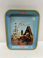 Front view of the 1957 Coca-Cola serving tray showing birdhouse, flowers, blossoming branch, and bottle graphics with blue background and yellow trim.
