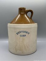 Front view of antique Kentucky Corn bourbon crock showing the conical brown glazed top, handle, and beige body with blue lettering.