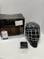 Photo of the Crown Royal Goalie Mask Ice Bucket out of the box, showing black goalie mask shaped ice bucket with silver cage bars and a black base stand, all on a white surface.
