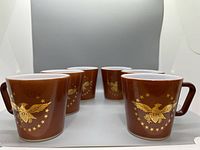 Front and side view of six brown Pyrex mugs arranged in a semicircle, showing gold eagle and star designs on each.
