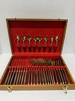 View of entire 32-piece cutlery set inside wooden box with red velvet lining. Small spoons held vertically in lid, forks and other utensils laid in bottom slots.