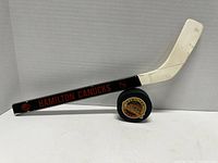 Side view of the souvenir hockey stick with 'Hamilton Canucks' text and logo on black shaft and white blade, the stick is balanced on the hockey puck.