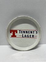 Front view of the round metal tray showing the Tennents Lager logo printed in white background with red and blue colors.
