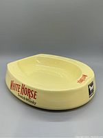 Side angle view showing cream-colored ceramic ash tray with red White Horse Scotch Whisky branding on the side and recessed ash area.