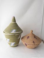 Front view of two lidded handwoven baskets, one green with white and blue designs, the other tan with brown diamond patterns, both with pointed lids.