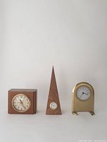 Photo of three clocks: a square wooden clock on left, a tall wooden pyramid shaped clock in middle, and a small brass finished clock on right.