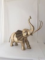 Front-side view of large brass elephant sculpture showing raised trunk, large curved ears, and solid body build on white backdrop.