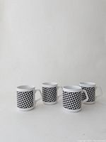 Four white porcelain mugs arranged together, showing the black and white houndstooth pattern on their sides and square-shaped handles.
