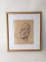 Pencil sketch portrait in gold frame with white mat, showing full framed artwork of elderly male profile on brown paper.